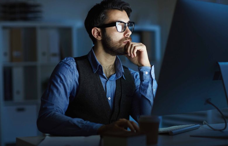 Man working at night Concentrated entrepreneur or manager sitting by desk in front of computer monitor while working at night